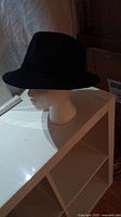 Black felt woman's hat displayed on a white mannequin head, showing side and front views, placed on white shelving unit.