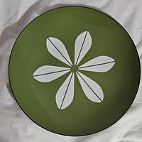 Top-down photo showing the full green enamel charger plate with white lotus flower design in the center, lying on a fabric surface.