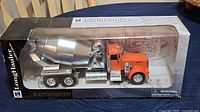 Photo of the LongHauler Kenworth W900 cement mixer truck model in its original black box packaging with clear window showing orange cab and silver mixer drum.