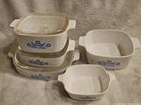 Five pieces of CorningWare Blue Cornflower bakeware arranged on a cloth backdrop, showing the design and shape differences between the dishes, including two with glass lids.