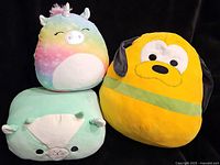 Three assorted Squishmallows plush toys including a pastel rainbow unicorn, a yellow dog, and a green alpaca on a black background.