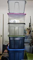 Stack of five assorted storage bins in various colors including gray, clear with pink lid, gray, dark blue, and blue.