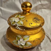 Front view of the vintage gold trinket dish showing hand-painted white flowers with green leaves and gold scroll designs. Lid features a round knob.