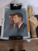 Poster featuring side-by-side portraits of JFK and RFK lying flat held by tape and stapler, alongside original mailing tube with tags attached.