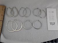 Photo showing seven pairs of silver hoop earrings on a light fabric background, ruler included for size reference. Includes round, oval, textured, and flat designs.