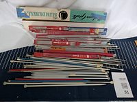 Multiple straight knitting needles in various sizes and colors laid flat, some in original packaging boxes