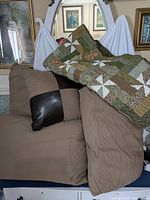 Four large taupe couch pillows, one with dark brown leather accent, small black throw pillow, and draped quilt with pinwheel pattern in greens, browns, and white.