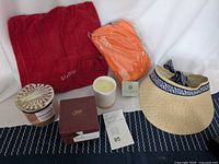 Full set arranged on fabric background showing red felt inflatable pillow, two scented candles including Soak surprise ring reveal and Saje natural candle, Aperol Spritz orange waist pack, and Sea and Grass woven summer visor.