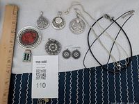 Full view of all jewelry laid out with ruler showing size for scale. Items include various silver pendants, matching earrings, silver chains, and black alternative necklaces.