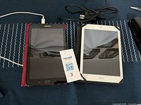 Two tablets placed side-by-side on a dark surface, showing Samsung tablet with red cover on the left and Acer tablet with white bezel on the right, with their charge cords above them.