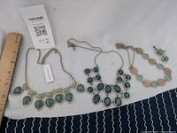 Photo of three gold-tone necklaces with teal and green stones and one pair of matching earrings laid out on white fabric alongside measuring ruler and tags.