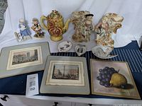 Photo of all lot items arranged on a surface including ceramic teapot, hot plates, ring holders, figurines, and framed prints.