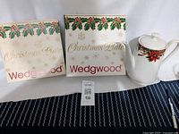 Photo of two Wedgwood Christmas plates in original boxes with holly and gold snowflake designs and one tall white poinsettia-patterned teapot.