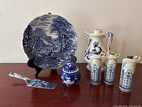 Wide shot showing the cake plate on stand, cake lifter, small ginger jar, three Delft bottles, and a jug bottle on wood surface.