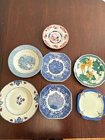 Seven assorted vintage and antique porcelain plates and dishes displayed on a wooden surface, showing varied decorative styles and colors including blue and white scenic prints, floral patterns, and oriental designs.