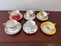 All six bone china teacups and matching saucers arranged on a wooden surface showcasing varied colors, shapes, and floral patterns