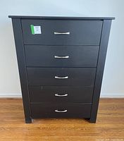Front view of black MDF wood dresser with 5 drawers and silver handles standing on wooden floor.