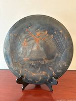 Full view of the round copper plate with incised Egyptian-style figures and floral decorations on a stand.