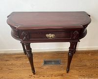 Front view of demilune mahogany side table with decorative carving and a single drawer with brass handle.