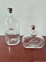 Two clear glass hand blown decanters by Erik Hoglund for Kosta Boda, standing side by side on a wood surface, showing front medallions and overall shape.