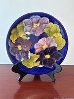 Front view of the Moorcroft plate showing Clematis flowers with pink, purple, and burgundy petals and green leaves against a cobalt blue background on a wooden table, displayed on a black stand.