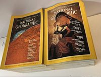 Photo of stacked National Geographic magazines with visible covers showing the December 1983 issue and another with a round object resembling a coin or medallion on cover.