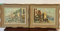 Photo of both matching oil paintings on canvas side-by-side showing coastal scenes with people and boats, each in beige carved wood frame.