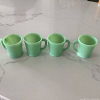 Four pale green Jadeite Fireking mugs with D handles on a white marble countertop shown from the front.