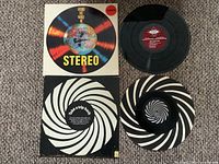 Photo showing two vinyl LP records with their respective album covers. One has a colorful circular design with 'Around the World in Stereo' text, the other displays a black and white spiral pattern with 'Take a trip back' text on the back cover.