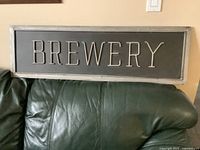 Front view of the metal brewery sign showing 'BREWERY' in raised metallic letters on a framed dark background resting on green leather sofa.
