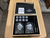 Top view of open wooden box showing two clear crystal whiskey glasses, eight grey whiskey stones arranged in foam slots, metal tongs with silicone tips, black cloth pouch with Sovyime logo, and instruction card.
