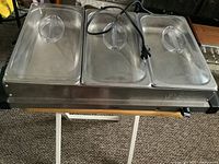 Photo showing the full triple compartment warming server, stainless steel body, clear domed lids, and attached power cord.