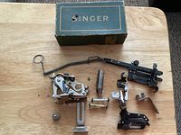 Top view of assorted Singer sewing machine attachments and presser feet arranged on wooden surface with original Singer box at rear.