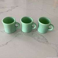 Three vintage jadeite mugs with C handles, positioned side by side on a marble countertop, showing front angle.