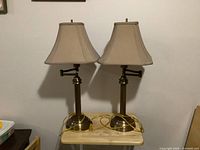 Two bronze coated table lamps with beige fabric shades placed side by side on a small table, showing overall condition and design.