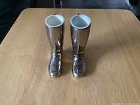 Photo shows a pair of silver plated miniature riding boot-shaped shot glasses placed on a wooden table, viewed from above