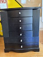 Front view of the black wooden jewelry box showing six drawers with white knobs and vertical groove detailing on sides.