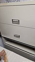 Front view of the beige metal filing cabinet showing metal drawers with recessed handles and open bottom drawer.