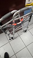 Side view of a folded silver aluminum folding walker with hand grips and red locking levers for height adjustment.