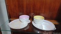 Two iridescent porcelain bowls with matching serving trays, one pink-rimmed and one yellow-rimmed, displayed on glass shelf inside wood cabinet.