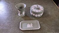 Three item set on table: green Wedgwood jasperware urn-shaped vase, covered round box with finial, and rectangular tray, each decorated with white relief scenes and floral border.