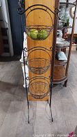 Full view of the 3 tier black wire fruit basket stand with three empty, round wire baskets attached vertically on a tall frame with scrollwork details.