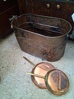 Photo showing a large hammered copper boiler with wire handles, placed on floor next to two copper pans with brass handles.