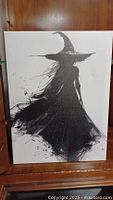 Rectangular 16 x 12 inch canvas with black and white silhouette of a witch wearing a pointed hat and flowing dress, styled with artistic brush strokes.