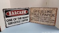 Two new metal signs with humorous sayings, each 8 inches by 12 inches, displayed against white backdrop.