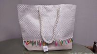 Front view of beige and white tote bag with colorful tassels on bottom edge, hanging rope handles.