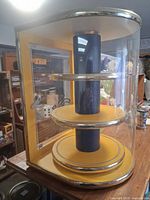 Full front and side view of the yellow and black retro spinner display case placed on a wooden surface surrounded by various collectibles and antiques.