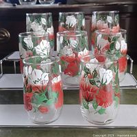 Photo of 8 clear juice glasses with midcentury floral designs in red, white and green, arranged in two rows on a reflective surface.