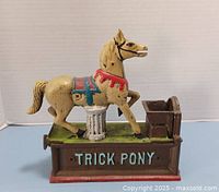 Front side of the Trick Pony cast metal bank, showing the horse figure atop a green base with the text 'TRICK PONY' in light blue on the front face. The horse is tan with red and blue saddle accents and positioned with one front leg raised.