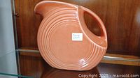 Side and angled views of vintage orange Fiestaware ceramic pitcher with concentric circle pattern and integrated handle displayed on a glass shelf.